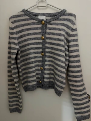 Randig kofta från H&M med guldknappar - Supermysig kofta från H&M i storlek XS, med grå och vita horisontella ränder. Koftan har fem snygga guldknappar framtill och är stickad i mjukt material. Perfekt för lager-på-lager och chill dagar. Långärmad och med rund hals.