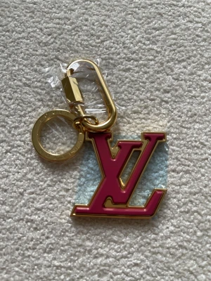  Vuitton rosa nyckelring - Säljer en snygg  Vuitton nyckelring med den klassiska -loggan i rosa och guld. Nyckelringen har en robust guldfärgad metallring och karbinhake, perfekt att hänga på väskan eller nycklarna. Lyxig och stilren accessoar som sticker ut.