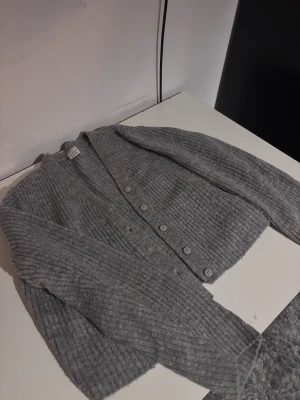 Grå croppad kofta med knappar - Säljer en grå ribbad kofta från Gina Tricot i croppad modell. Koftan har långa ärmar, v-ringad hals och knappar framtill. Perfekt att slänga över ett linne eller t-shirt för en chill look. Mjuk stickad känsla och snygg passform.