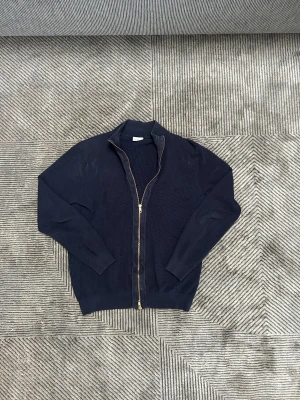 Marinblå helzip från John Henric - Snygg marinblå helzip från John Henric i storlek M. Tröjan har ett ribbat mönster, hel dragkedja framtill och hög krage. Materialet är stickad bomull som känns mjukt och skönt mot huden. Perfekt för dig som gillar stilrena och klassiska plagg.