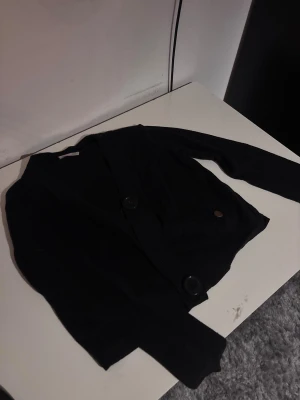 Svart croppad kofta med stora knappar - Snygg svart croppad kofta med långa ärmar och två stora knappar framtill. Koftan har en enkel och stilren design som är lätt att matcha med olika outfits. Materialet känns mjukt och stretchigt, perfekt för dig som gillar en bekväm men trendig look.