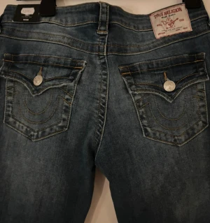 Helt nya True  Religion med lapp! - Helt nya true religion! Kappan och allt sitter kvar! Aldrig använda! Snygga blå jeans från True Religion, modell Joey LR Flare Flap. Låg midja och utställda ben med klassiska fickor bak och silverfärgade knappar. Tuffa kontrastsömmar och slitningar ger en cool vibe. Perfekt för dig som gillar statement-jeans med unik design.