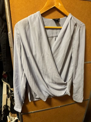 Ljusgrå/ blå omlottblus från Lindex - Säljer en ljusgrå/blå omlottblus från Lindex i storlek 36. Blusen har långa ärmar och ett snyggt omlottfall framtill som ger en cool och avslappnad look. Materialet är mjukt och tunt, perfekt för att styla med jeans eller kjol.