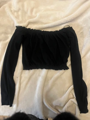 Svart offshoulder croppad topp - Säljer en svart offshoulder topp med långa ärmar och croppad passform. Toppen har en lätt rynkad design vid hals och nederkant som ger en cool och trendig look. Perfekt för dig som gillar att sticka ut med en stilren vibe.
