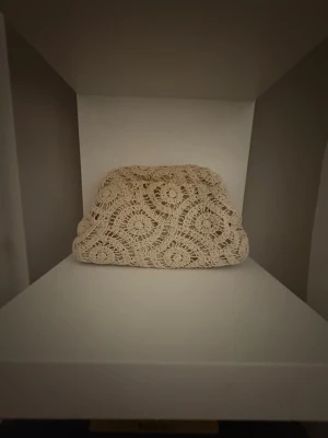 Beige virkad kuvertväska - Superfin beige kuvertväska med virkat mönster och snyggt innerfoder. Väskan har ett unikt cirkelmönster och stängs med ett klassiskt spänne. Perfekt för dig som vill sticka ut med en bohemisk touch. lite slitning där inne som är ett hål men inget som märks på utsidan!