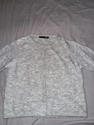 Ljusgrå tröja från 157 med pärlknappar - Supermjuk ljusgrå tröja från 157 med rund hals och snygga pärlknappar framtill. Tröjan är stickad och har kort ärm, perfekt att slänga på sig när det är kyligt. Enkel och stilren design som funkar till det mesta. Använd ca 2-3 gånger