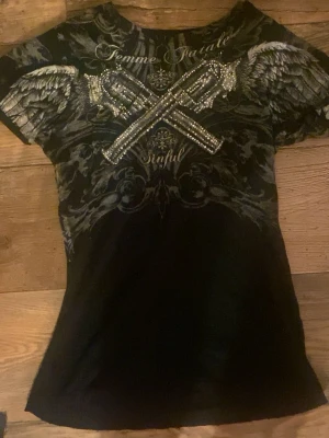 Svart Sinful t-shirt med vingar och rhinestones - Svart t-shirt från Sinful med coolt tryck av vingar och korslagda pistoler på framsidan, dekorerad med rhinestones. Baksidan har ett stort kors och detaljerade mönster. T-shirten har korta ärmar och är gjord i mjuk bomull. Små hål runt halsen och sen ett litet hål med små små hål brövid längst ner på tröjan på framsidan men inget jag tycker märks när man använder den. En av mina favorit tröjor som alltid gör outfitten fire. 🔥