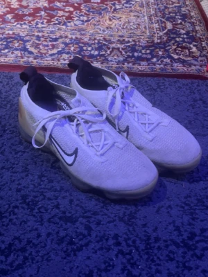 Nike Vapormax vita sneakers - Nike Vapormax sneakers i vit färg med svart swoosh på sidan. Skorna har en stickad ovandel och genomskinlig platt sula med unik bubbeldesign. Snörning framtill och svarta detaljer vid hälen. Perfekta för dig som gillar sportig och modern stil.