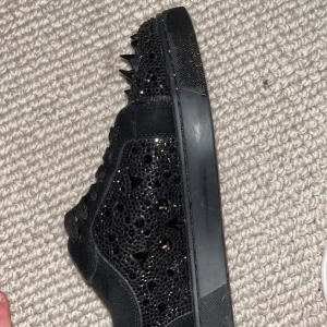 Christian Louboutin svarta sneakers med nitar - Säljer ett par svarta sneakers från Christian Louboutin med ikonisk röd sula skorna saknar även bara en nit . Skorna har snörning, rund tå och är täckta med svarta nitar och sten detaljer på sidorna och tån. Materialet är mocka och skinn, vilket ger en lyxig vibe. 