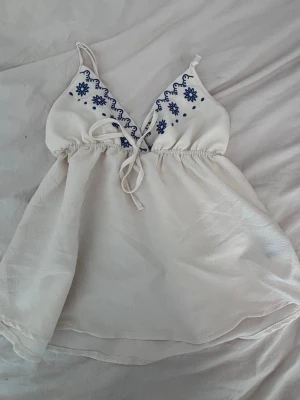 Vit broderad linnetopp med snörning - Säljer en söt vit linnetopp med blå broderade blommor vid bysten och snörning framtill. Toppen har tunna axelband och är lätt och luftig med en flowy passform. Perfekt för varma dagar och ger en fräsch sommarkänsla.