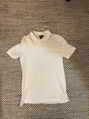 Vit slim fit piké från H&M - Vit piké från H&M i slim fit-modell, storlek S. Klassisk pikékrage med två knappar och diskret svart kant på kragen. Tillverkad i 100% bomull för skön känsla och snygg passform. Perfekt för en clean och fräsch look.