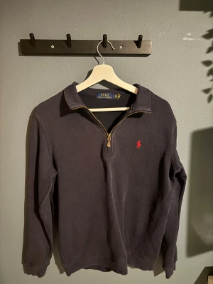 Mörkblå half zip tröja Polo Ralph Lauren - Mörkblå långärmad tröja från Polo Ralph Lauren med klassisk röd broderad logga på bröstet. Tröjan har en snygg krage och half zip-dragkedja i guldton. Tillverkad i mjuk bomull, perfekt för chill höst- och vinterdagar.