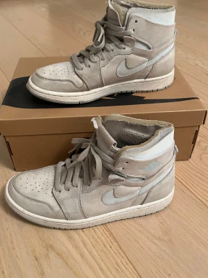 Nike Jordan 1 High  - Nike Jordan 1 High sneakers i ljusgrå mocka med vita detaljer och klassisk Swoosh på sidan. Skorna har snörning, perforerad tå och högt skaft för en ikonisk look. Perfekt för dig som gillar streetwear och vill sticka ut med stilrena kicks.