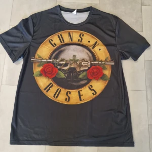 Guns N' Roses svart t-shirt - Svart t-shirt med stort Guns N' Roses-tryck både fram och bak. Trycket har gul cirkel, röda rosor och metalliska revolvrar. Rund hals och korta ärmar. Materialet känns lätt och syntetiskt, perfekt för dig som vill sticka ut med en ikonisk bandtröja.