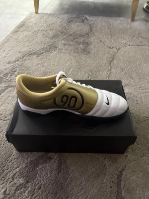 (R9) Nike Total 90 III fotbollsskor guld/vit - Nike Total 90 III fotbollsskor med snygg guld och vit färgkombination, svart 90-logga på sidan och klassisk Swoosh. Skorna har snörning, rund tå och platt sula. Tillverkade i syntetmaterial med skinnkänsla för bra komfort och passform. Kan alltid förhandla!