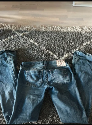 True Religion blå bootcut jeans - Säljer ett par snygga blå jeans från True Religion i klassisk bootcut-modell. Jeansen har ikoniska bakfickor med broderad logga, silverfärgade knappar och kontrastsömmar. Inrebenslängd 76