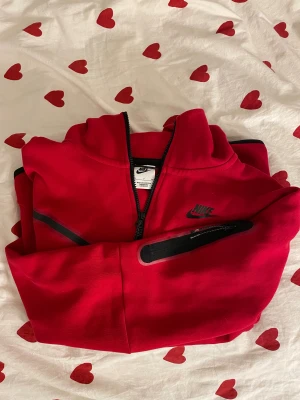 Röd Nike tech fleece - Säljer en röd Nike tech fleecemed svart logga på bröstet och svarta detaljer. Tröjan har hel dragkedja framtill, huva och en ficka med dragkedja på ärmen. Mycke bra skick!
