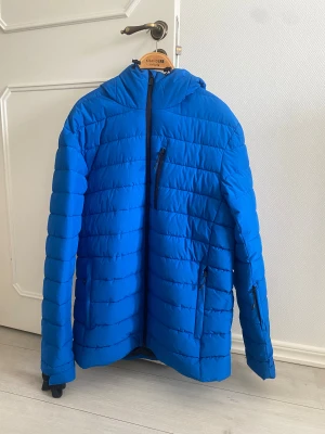 Blå McKinley pufferjacka med huva - Säljer en blå pufferjacka från McKinley i storlek Large. Jackan har huva, flera dragkedjeförsedda fickor och quiltad design. Perfekt för kalla dagar och har en sportig look med snygga svarta detaljer vid dragkedjorna.