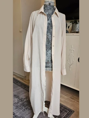 Ljusbeige långskjortklänning - Lång skjortklänning i ljusbeige med klassisk krage och knappar hela vägen framtill. Tillverkad i ett mjukt, strukturerat bomullstyg. Långa ärmar med manschetter. 