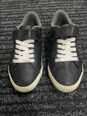 Sneakers - Svarta och vita sneakers från H&M med tryck av gul grävmaskin på sidan. Skorna har vita snören, kardborreband och grått foder. Baktill finns en gul hängare. Skorna är unisex, i storlek 33 och inte jättemycket använda