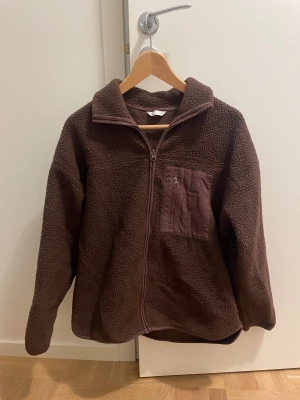 Brun teddyjacka - Mysig brun teddyjacka från Cubus med hel dragkedja framtill och en praktisk bröstficka med zip. Jackan har hög krage och långärmade ärmar, perfekt för kyliga dagar. Materialet är fluffigt fleece som ger en skön och avslappnad vibe.