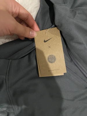 Grå Nike PSG träningsset herr S - Säljer ett grått träningsset från Nike x Paris Saint-Germain med rosa PSG-logga och rosa swoosh-detaljer. Setet består av långärmad tröja med mönstrade ärmar och matchande byxor med dragkedja vid benslut. Materialet är syntet och passar perfekt för fotbollsträning.