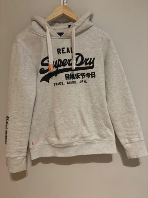 Superdry Hoodie Grå - En stilren Superdry hoodie i färgen grå med svart tryck. Den är knapp använd och är i superbra skick! Det står storlek M i den men sitter mer som storlek S. Pris kan diskuteras! 