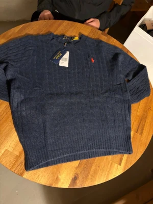 Marinblå stickad tröja Polo Ralph Lauren - Snygg marinblå stickad tröja från Polo Ralph Lauren med diskret röd logobroderi på bröstet. Tröjan har ribbade muddar och rund hals, perfekt för dig som gillar klassisk stil med en modern touch.
