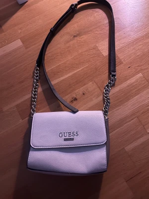 Guess väska  - Snygg vit väska från Guess med silverfärgad kedja och justerbar axelrem. Insidan har ett fack med dragkedja och Guess-logga och en korthållare. Perfekt för dig som gillar clean och modern stil. Änvänd fåtal gånger! 🩷