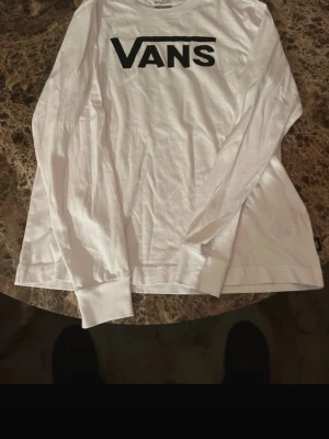 Vit långärmad topp från Vans - Säljer en vit långärmad topp från Vans med stort svart logotryck på bröstet. Toppen har rund hals och breda muddar vid ärmsluten. Perfekt för dig som gillar streetwear och enkel stil.