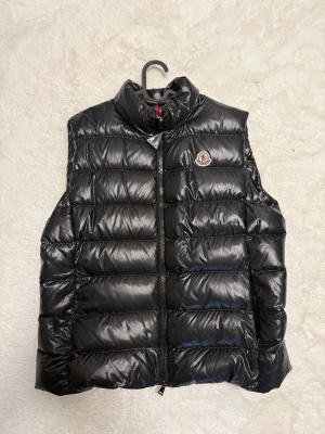 Svart dunväst från Moncler - Snygg svart dunväst från Moncler med quiltad design och hög krage. Västen har dragkedja framtill och Moncler-logga på bröstet. Materialet är glansigt och känns lyxigt, perfekt för att addera lite edge till din outfit. Passar dig som gillar stilren och sportig look.