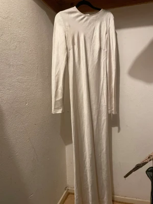 Beige  långärmad maxiklänning - En stilren, beige långklänning med lång ärm och rund halsringning. Klänningen har en rak och enkel siluett, dragkedja i ryggen och är tillverkad i ett slätt tyg som ger en elegant känsla. Perfekt för dig som gillar minimalistisk och clean stil.