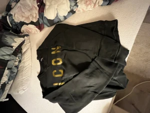 Svart sweatshirt DSQUARED2 ICON S - Svart sweatshirt från DSQUARED2 i storlek S med guldfärgad text 'ICON' på framsidan och DSQUARED2-logga i guld på ryggen. Rund halsringning med snygga sömdetaljer. Mjuk insida, perfekt för chill och streetwear. Materialet är bomullsmix. Använd kanske två gånger, passformen var inte för mig.