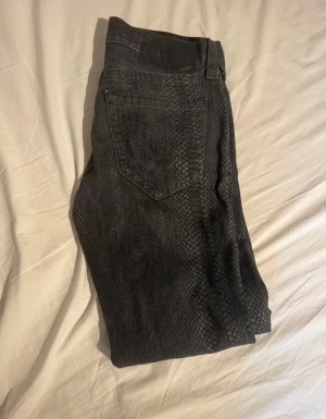 True Religion svarta jeans med ormmönster - Säljer ett par svarta jeans från True Religion med unikt ormmönster över hela byxan. Modellen är rak och har klassisk femficksdesign samt läderpatch bak i midjan. Materialet är jeans och passformen är rak, perfekt för dig som vill sticka ut.