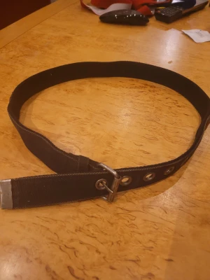 Svart nitbälte med metallhål - Svart bälte med silverfärgade nitar och metallhål längs hela bandet. Bältet har en enkel metallspänne och är gjort i slitstarkt syntetmaterial. Perfekt för att ge din outfit en edgy vibe. Passar till jeans eller över en skjorta för att bryta av stilen.