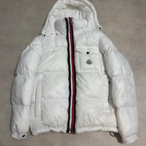 Vit Moncler pufferjacka med huva - Säljer en fet vit pufferjacka från Moncler med huva och coolt Moncler-märke på bröstet. Jackan har en röd, vit och blå rand längs dragkedjan, stora puffiga sektioner och en ficka med knapp framtill. Insidan har en comic strip-etikett och DIST-certifiering . 