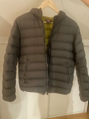 Grå Colmar pufferjacka med huva - Säljer en grå pufferjacka från Colmar med huva och två sidofickor med dragkedja. Jackan har en mörkgrön insida och är quiltad med breda kanaler. Perfekt för dig som vill ha en clean och stilren look under höst och vinter.
