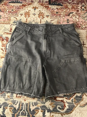 Grå denimshorts från Zara, strl 42 - Säljer ett par grå denimshorts från Zara i storlek 42. De har råa kanter, stora fickor både fram och bak samt Blend-märke på sidan. Klassisk femficksmodell med bälteshällor och knappstängning. Perfekt för dig som gillar street och chill vibe.