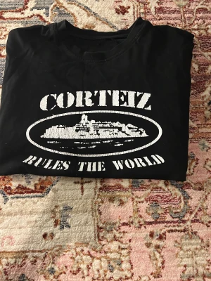 Svart Corteiz t-shirt med tryck - Svart t-shirt från Corteiz med stort vitt tryck på både fram- och baksidan. Framsidan har texten 'Corteiz Rules The World' och ett grafiskt motiv, medan baksidan har en stor textdesign. Klassisk rund hals och korta ärmar, perfekt för streetwear-stilen.