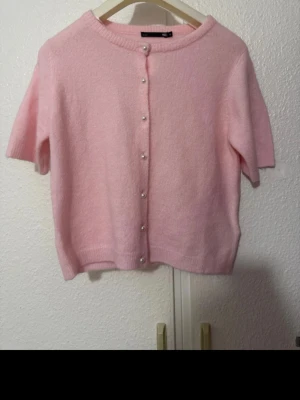 Ljusrosa kofta med pärlknappar - Supersöt ljusrosa kofta med korta ärmar och pärlknappar framtill. Mjuk och fluffig känsla, perfekt för dig som gillar en gullig och trendig look. Passar till både jeans och kjol för en söt outfit.