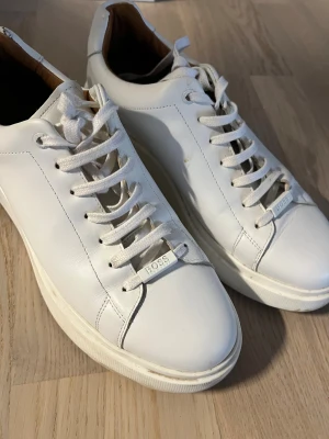 Vita sneakers från BOSS Bulton i skinn - Ordinarie pris 2800kr. Snygga vita sneakers från BOSS med stilren design och rund tå. Skorna är tillverkade i slätt skinn och har vita snören samt platt sula med BOSS-logga på sidan och snörningen. Perfekta för dig som gillar minimalistisk och clean stil.