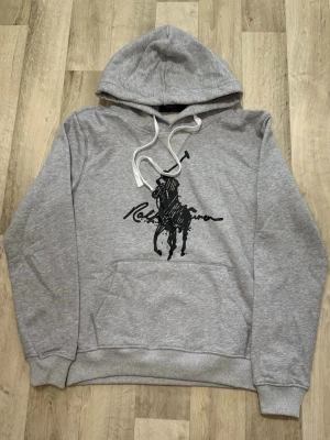 Grå hoodie – Polo Ralph Lauren  - Snygg och bekväm grå hoodie med stor Polo Ralph Lauren-logga på bröstet. Mjuk bomullskänsla, justerbar huva och känguruficka fram. Perfekt för vardag, skola eller träning.