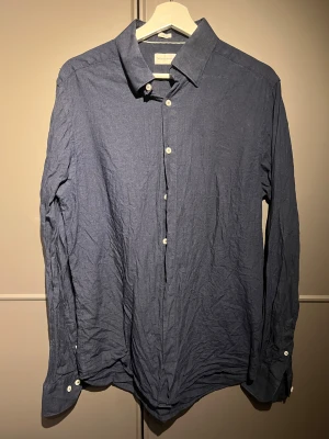 Mörkblå skjorta These Glory Days L - Snygg mörkblå skjorta från These Glory Days i storlek L, slim fit. Skjortan har klassisk krage, vita knappar och långa ärmar. Perfekt för dig som gillar stilren och enkel design. Tillverkad i bomull för en skön känsla hela dagen.