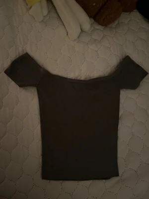 Mörkgrå ribbad offshoulder topp - Säljer en mörkgrå ribbad topp med offshoulder-design och korta ärmar. Snygg, tight passform som framhäver axlarna. Perfekt att styla med jeans eller kjol för en trendig look. Finns liten fläck på tröjan
