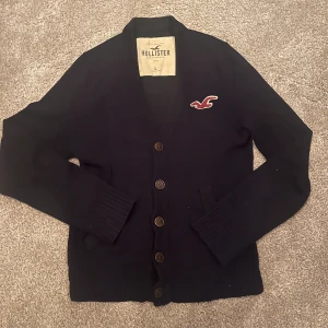 Mörkblå kofta från Hollister - Snygg mörkblå kofta från Hollister med bruna knappar och broderad logga på bröstet. Koftan har ribbade muddar, två fickor framtill och är långärmad. Perfekt för dig som gillar klassisk och chill stil.