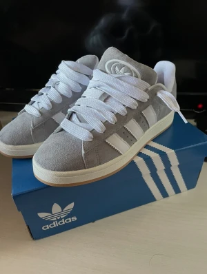 Adidas Campus 00s - Snygga Adidas Campus sneakers i grå mocka med vita detaljer och klassiska tre ränder på sidan. Skorna har vit sula, rund tå och breda vita skosnören. Adidas logga på plös och häl. Perfekta för dig som gillar stilrena och tidlösa sneakers.