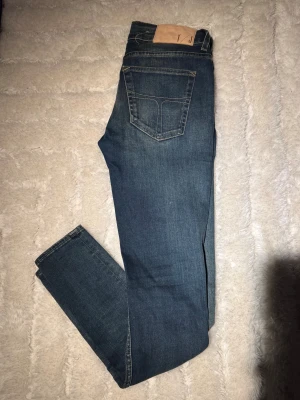 Blå jeansbyxor från Tiger Of Sweden, 30/32 - Snygga blå jeansbyxor från Tiger Of Sweden, tillverkade i Italien. Klassisk femficksmodell med raka ben och mörk tvätt. Skinnpatch med logga baktill i midjan. Perfekt passform för dig som gillar stilrena och tidlösa jeans. W30, L 32