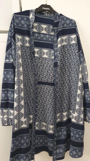 Klänning - Säljer en snygg långärmad klänning från Boohoo i storlek 36. Klänningen har ett unikt blåvitt mönster med olika geometriska och blommiga detaljer. Den är rak i modellen och har en skön passform som känns både chill och stilren.