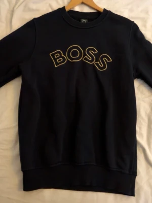 Svart/mörkblå sweatshirt från BOSS med gul text - Svart/mörkblå sweatshirt från BOSS i storlek S med gul broderad BOSS-logga på bröstet. Tröjan har rund hals, ribbade muddar och är långärmad. Materialet är mjukt och känns som bomull, perfekt för chill dagar. Enkel och stilren design som funkar till jeans eller joggers.