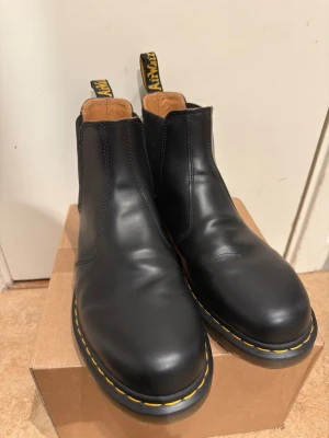 Dr. Martens 2976 YS Chelsea boots svart - Helt oanvända. Säljer ett par svarta Dr. Martens Chelsea boots i storlek EU48 med den ikoniska gula sömmen runt sulan. Tillverkade i skinn med AirWair-bounce sula för extra komfort. Lite creasing där foten böjs på ovansidan.